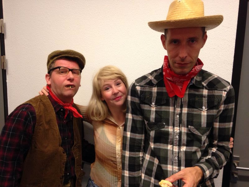 Drie personen. Een is verkleed als boer, een blonde vrouw lacht vriendelijk en de derde is verkleed als cowboy.