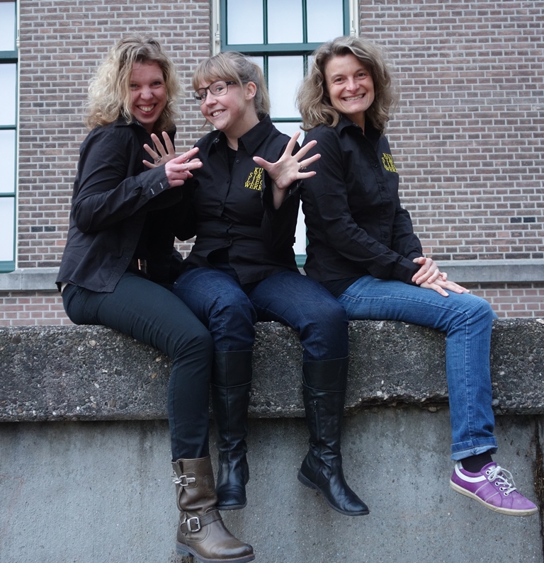 Drie vrouwen zitten op een muurtje met hun benen erover heen. Ze lachen. Ze hebben zwarte blouses aan met het Kunst & Vliegwerk logo.