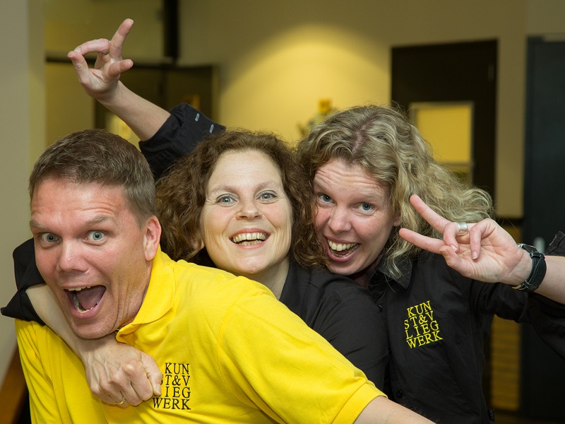 Een man in een geel t-shirt die met open mond lacht en twee vrouwen in zwarte blouse erachter lachen ook en maken beide een v-teken met hun vingers.
